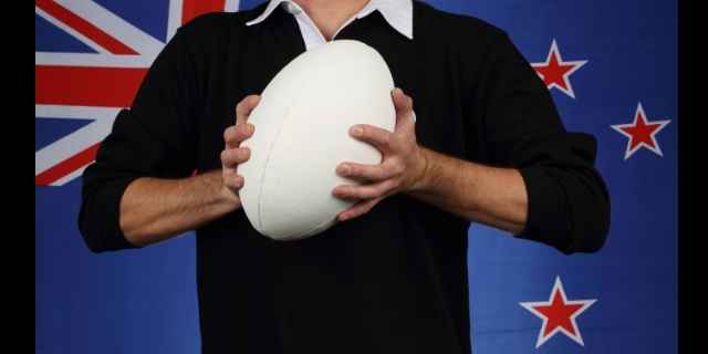 player in all black shirt holding a ball in front of a new zealand flag